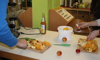 Zubereitung eines Obstsalates auf einem Küchentisch. Das Obst wird von 2 Personen auf Holzbrettern klein geschnitten.