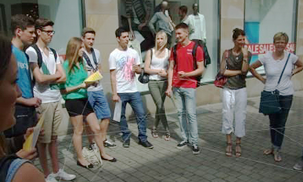 Eine Gruppe Jugendliche steht im Kreis auf einer Einkaufsstraße vor den Schaufenstern eines Modegeschäfts.
